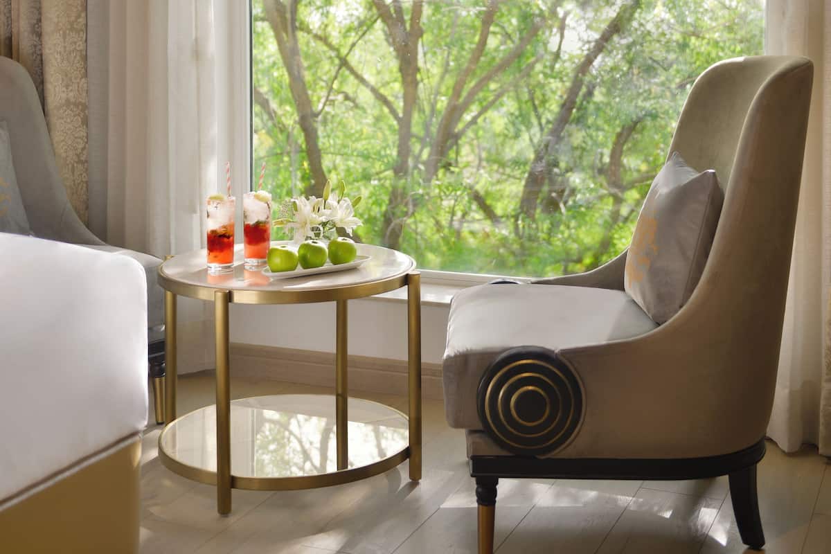 A hotel room with a bed, a chair, a small table with drinks and flowers, and a large window with a view of trees.