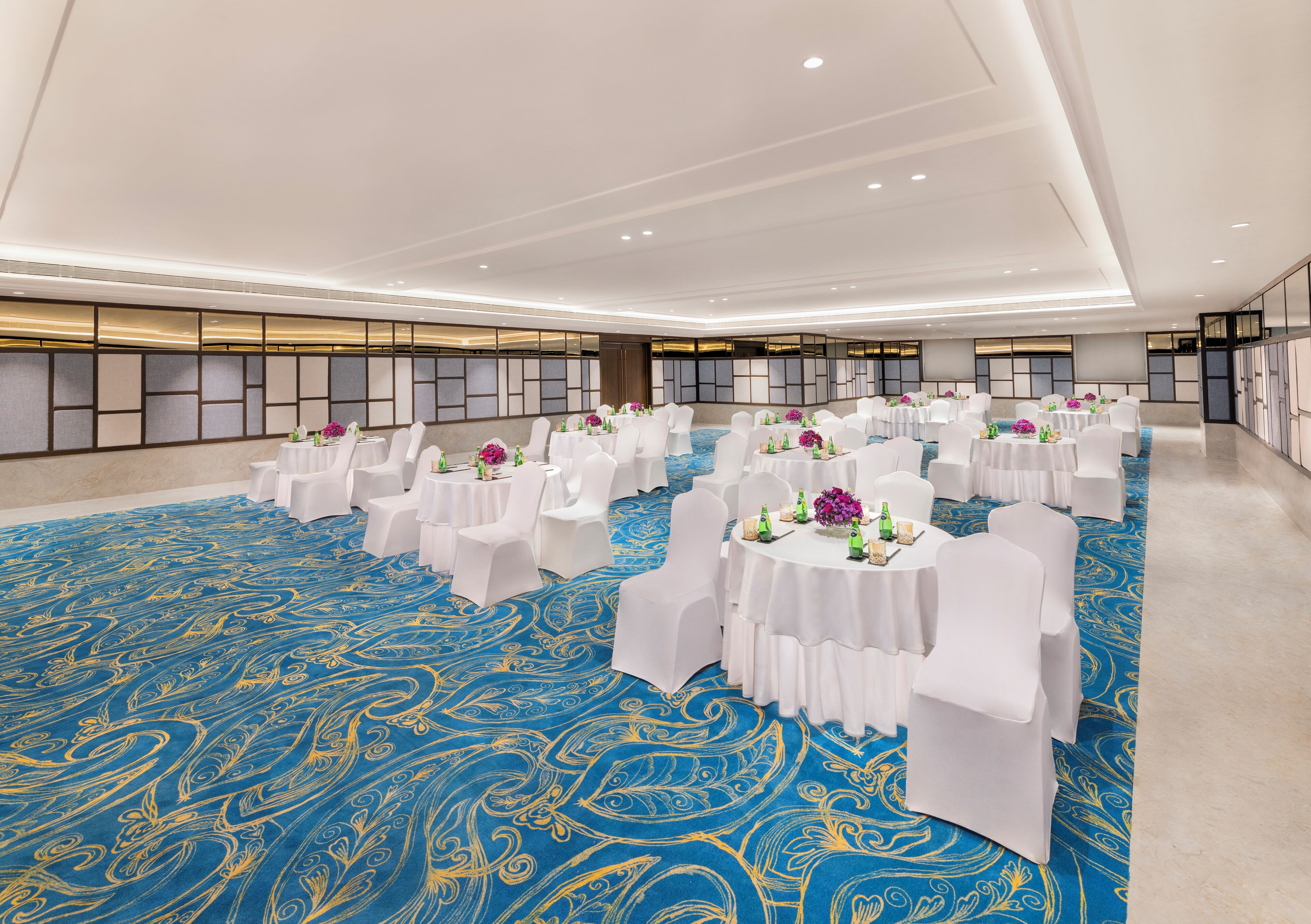 Un amplio salón de banquetos bien iluminado, con mesas redondas y sillas blancas, que presenta una alfombra con estampado azul y un diseño de techo moderno.