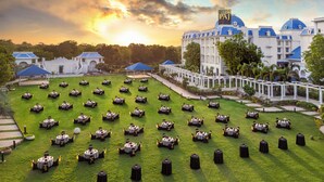 A formal outdoor dining area with multiple round tables arranged on a lawn, surrounded by white columns and a pavilion. - Taj Gandhinagar Resort & Spa (Mansa)