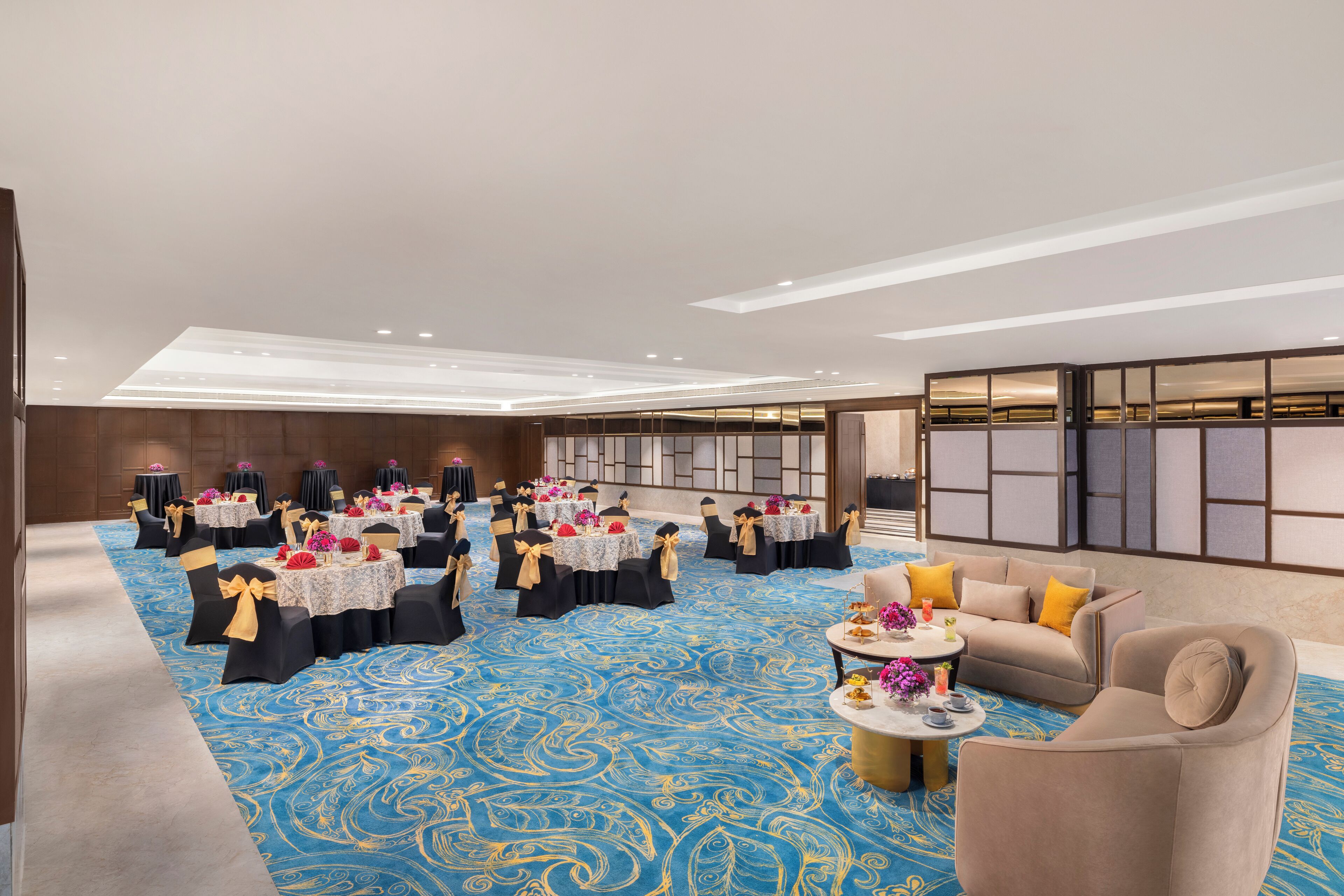 A spacious room with a blue patterned carpet, multiple round tables with white tablecloths and black chairs, and a sofa with yellow cushions.