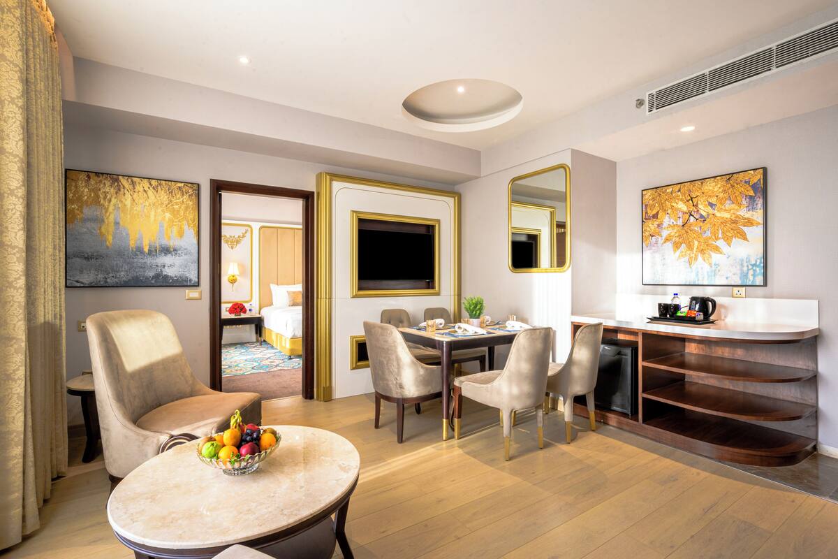 A modern hotel room with a dining area, a sitting area with a fruit bowl, and a workspace with a desk and chair.