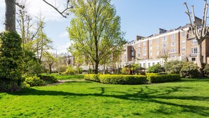 A well-maintained garden with a gazebo, neatly trimmed hedges, and a pathway leading to a row of brick townhouses. - Light Show, by Veeve (London)