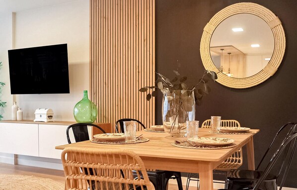 Un comedor con una mesa de madera preparada para cuatro personas, rodeada de sillas de mimbre. Un espejo grande cuelga en la pared, y un televisor de pantalla plana está montado sobre la mesa.