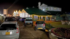 Un estacionamiento con varios coches, un edificio con amplias ventanas y un toldo verde, y un cartel con texto.