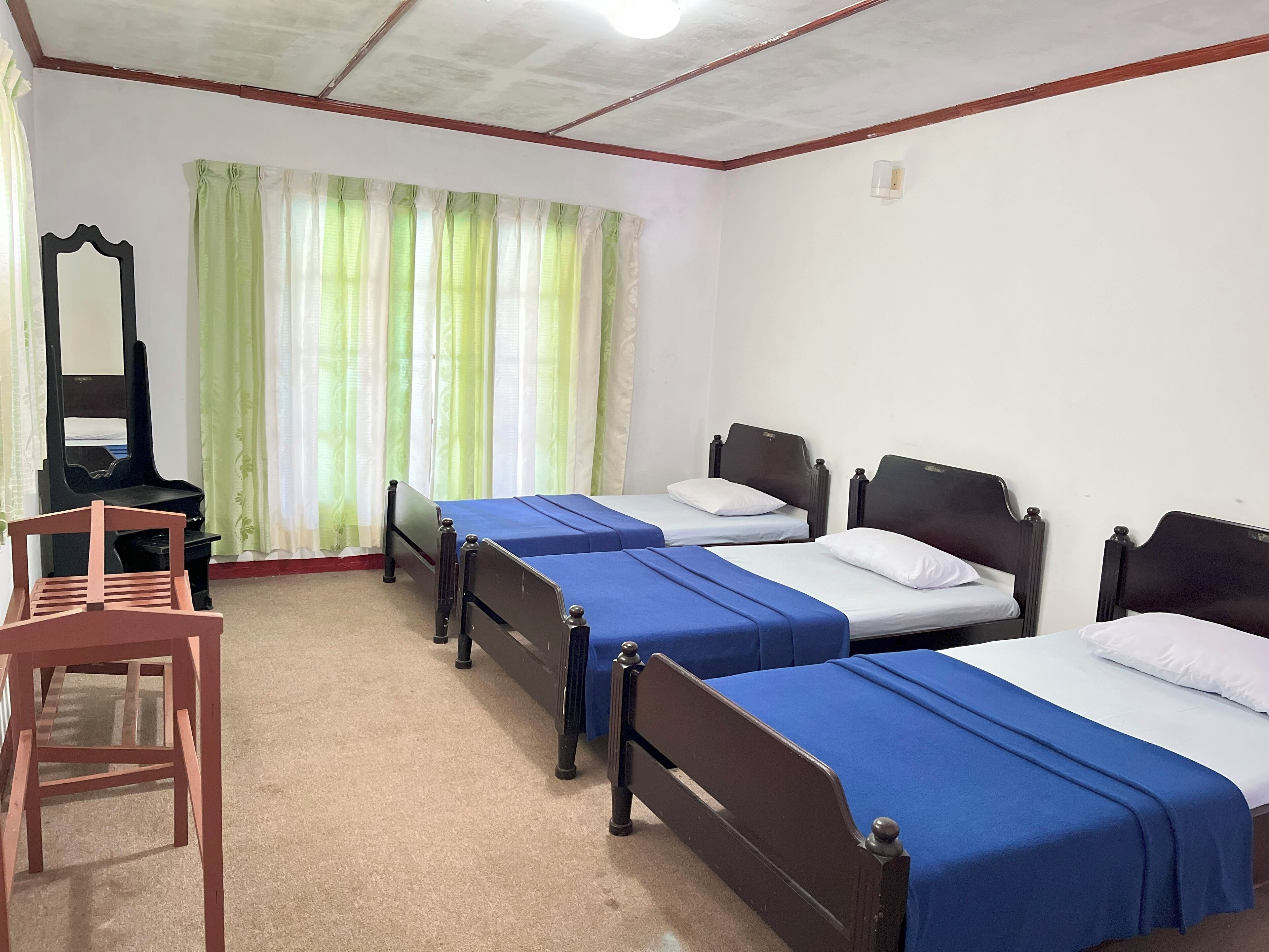 A room with three single beds, a wooden chair, a mirror, and a window with green curtains.
