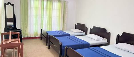 A room with three single beds, a wooden chair, a mirror, and a window with green curtains.