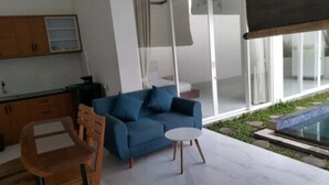 Una sala de estar moderna con un sofá azul, una silla de madera y una mesita blanca. Hay un ventanal que da a un balcón con piscina.