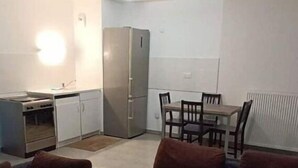 Een woonkamer met een zithoek, een eettafel met stoelen, een keuken met een koelkast, en een klein witte tafeltje.