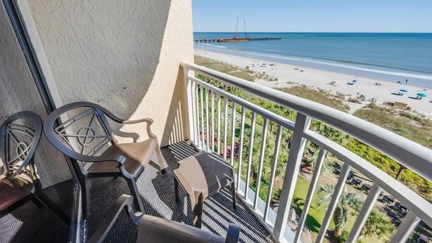 Un balcon avec deux chaises et une petite table, donnant sur une plage avec une jetée et des parasols.