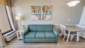 Un salon avec un canapé de couleur turquoise, une petite table d’appoint avec une lampe, une table à manger blanche avec des chaises, et deux œuvres d’art mural représentant des palmiers.