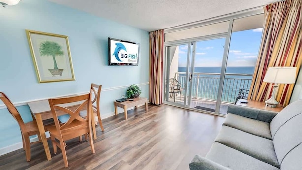 Una habitación con una mesa de comedor de madera, sillas, un sofá, un televisor y un balcón con vistas al océano.