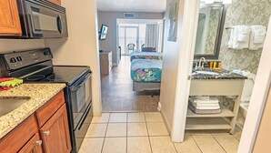 A compact hotel room with a kitchenette, a bed with a floral bedspread, a bathroom with a sink and mirror, and a view of the outside. - Direct Oceanfront Amazing Views In Camelot Resort! (Myrtle Beach)