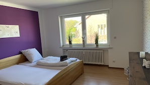 A bedroom with a wooden bed, white bedding, a purple accent wall, a window with plants, and a radiator. - Apartment Geronimo (Werne)