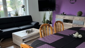 A modern living room with a black sofa, wooden chairs, a dining table with purple placemats, a flat-screen TV, and a purple accent wall. - Apartment Geronimo (Werne)