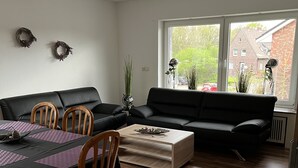 A living room with a black sofa, wooden dining table, and chairs. There is a window with a view of buildings and greenery outside. - Apartment Geronimo (Werne)