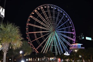 A large Ferris wheel with multiple cabins, illuminated with colorful lights, and a building with a sign that reads "SkyWheel" in the foreground. - Sand Dunes Resort 7th Floor Direct Oceanfront Pool (Myrtle Beach)