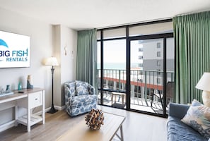A modern living room with a sofa, a coffee table, and a desk. There is a large window with curtains, a view of the sea, and a sign for 'BIG FISH RENTALS'.