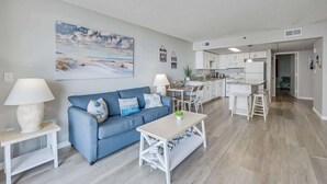 A modern living room with a blue sofa, a white coffee table, and a kitchen in the background. - Sand Dunes Resort 7th Floor Direct Oceanfront Pool (Myrtle Beach)