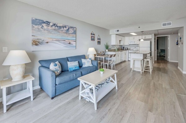 A modern living room with a blue sofa, a white coffee table, and a kitchen in the background.