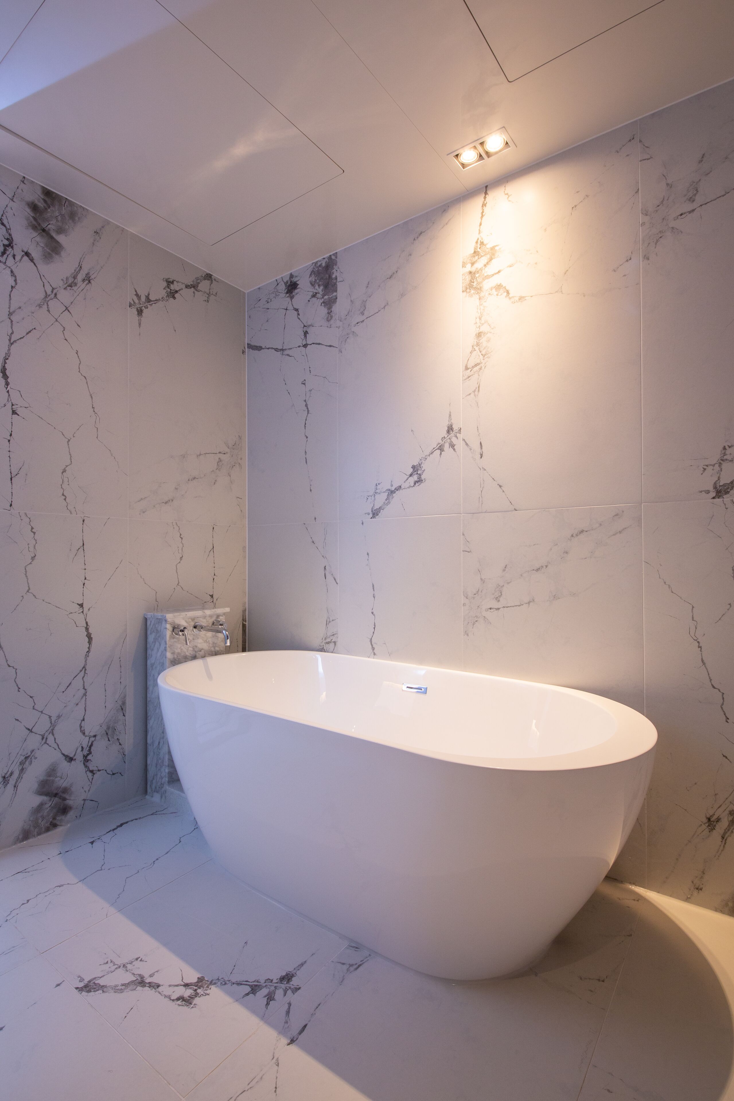 A modern bathroom with a freestanding bathtub, marble walls, and recessed ceiling lighting.