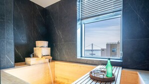 A modern bathroom with a wooden bathtub, a window with a view of a bridge, and a wooden bench with a green bottle and glasses. - Gwanganri The Club hotel (Busan)