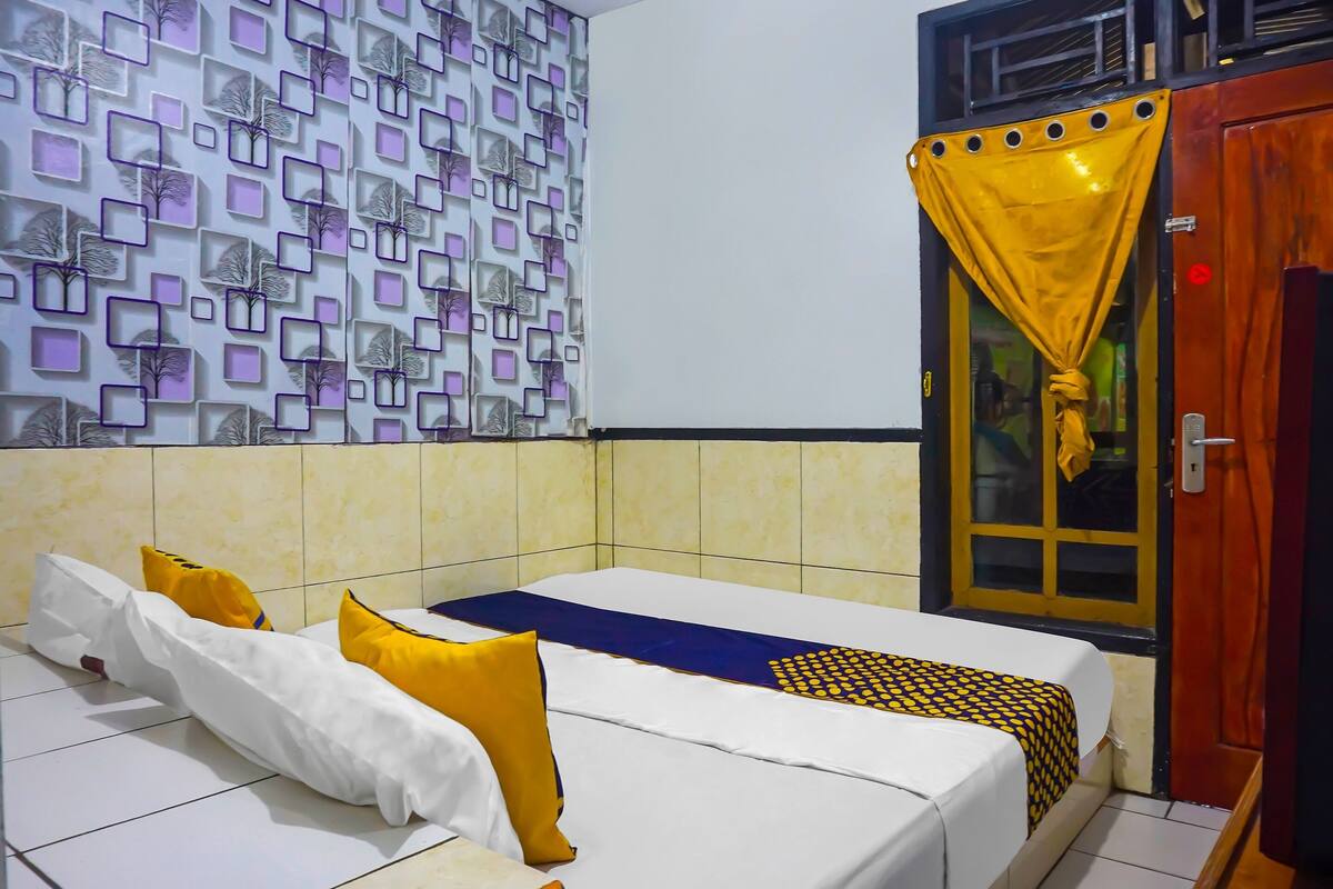 A hotel room with a bed, a patterned wall, a window with yellow curtains, and a door.