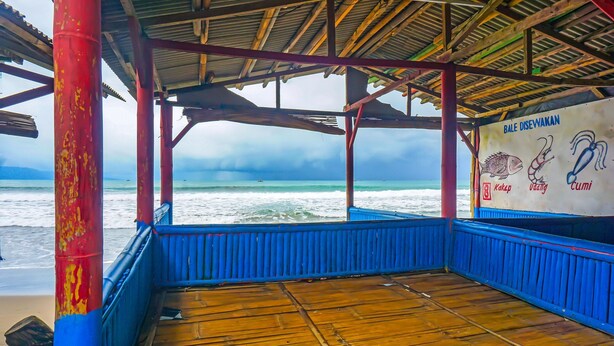 Una terraza de madera cubierta con vista al océano, un letrero que anuncia mariscos locales y un pilar rojo.