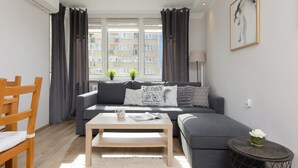 A modern living room with a grey sofa, a wooden coffee table, and a view of a building through large windows. - Comfortable Apartment in the Center | Balcony and Air Conditioning (Warszawa)
