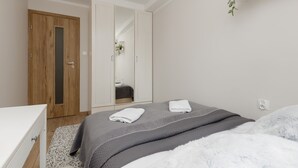 A bedroom with a wooden wardrobe, a bed with grey bedding, a white bedside table, and a hanging plant. - Comfortable Apartment in the Center | Balcony and Air Conditioning (Warszawa)