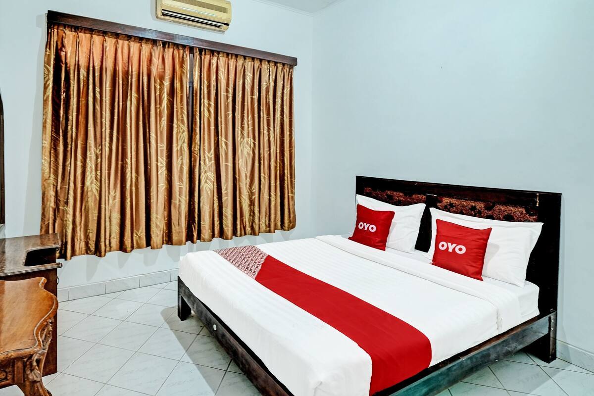 A neatly made bed with white and red bedding, a wooden headboard, and a window with brown curtains.