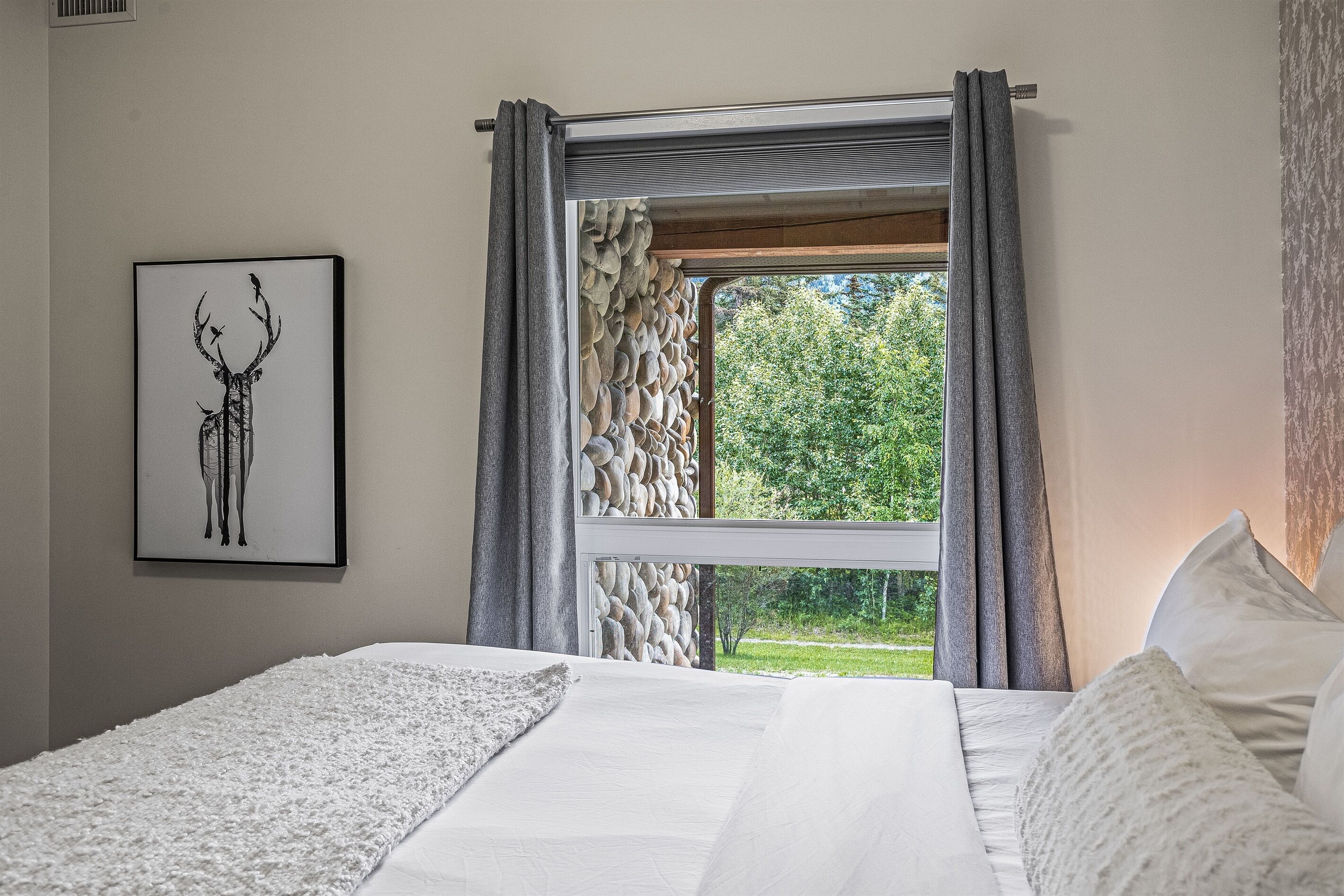 A bedroom with a large bed, a framed deer artwork, and a window with a stone wall view.