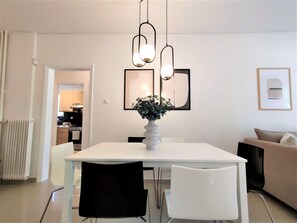 Un espace repas moderne avec une table blanche, des chaises noires et blanches, un vase contenant des plantes vertes et une œuvre d’art abstraite accrochée au mur.