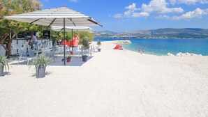 A beachside café with outdoor seating and umbrellas, overlooking a clear blue sea and distant hills. - A3 apt With Balcony and sea View, 3 min to Beach (Trogir)