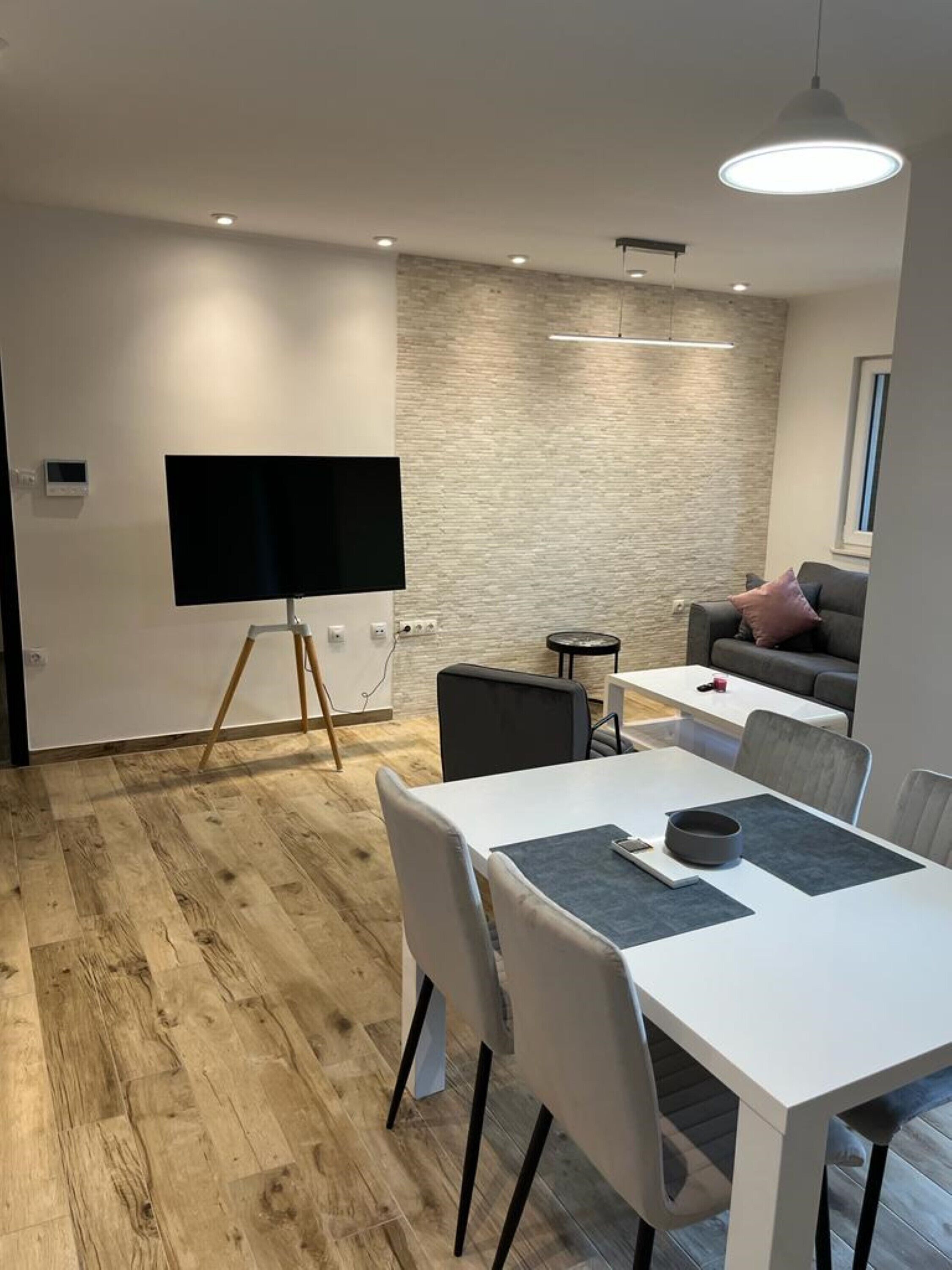 A modern living room with a flat-screen TV on a stand, a dining area with a white table and chairs, and a grey sofa.