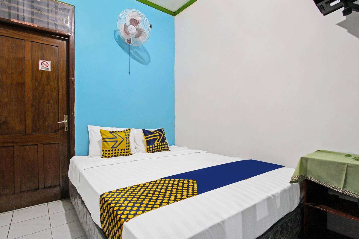 Ein Schlafzimmer mit Holztür, einem Bett mit weißer Bettwäsche und einer blau-gelb gemusterten Tagesdecke, zwei Kissen mit geometrischen Mustern und einem Deckenventilator.