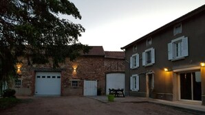 A courtyard with a dirt ground, two garage doors, a stone building, and a modern building with windows and lights. - Chez Arnaud (Saint-Jean-Saint-Maurice-sur-Loire)
