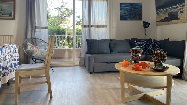 A living room with a sofa, a dining table, a chair, and a balcony with curtains. - Appartement rez de Chaussée (Mandelieu-la-Napoule)