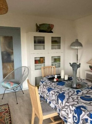 Un comedor con una mesa redonda cubierta con un mantel floral, rodeada de sillas. Encima de la mesa, hay un aparador con puertas de vidrio, y una lámpara de pie a la derecha.