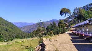 Une rangée de cabanes, reliées par un chemin de terre, est entourée de verdure et s’étend au pied de montagnes qui se dressent à l’arrière-plan.