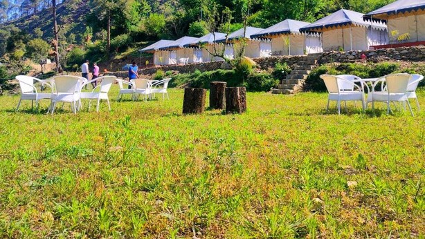 Une série de tentes avec des chaises blanches, installées dans un champ herbeux, le tout sur fond de colline boisée.