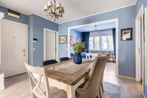 Un coin repas avec une table en bois, des chaises et un lustre. On aperçoit un salon avec un canapé et une fenêtre.