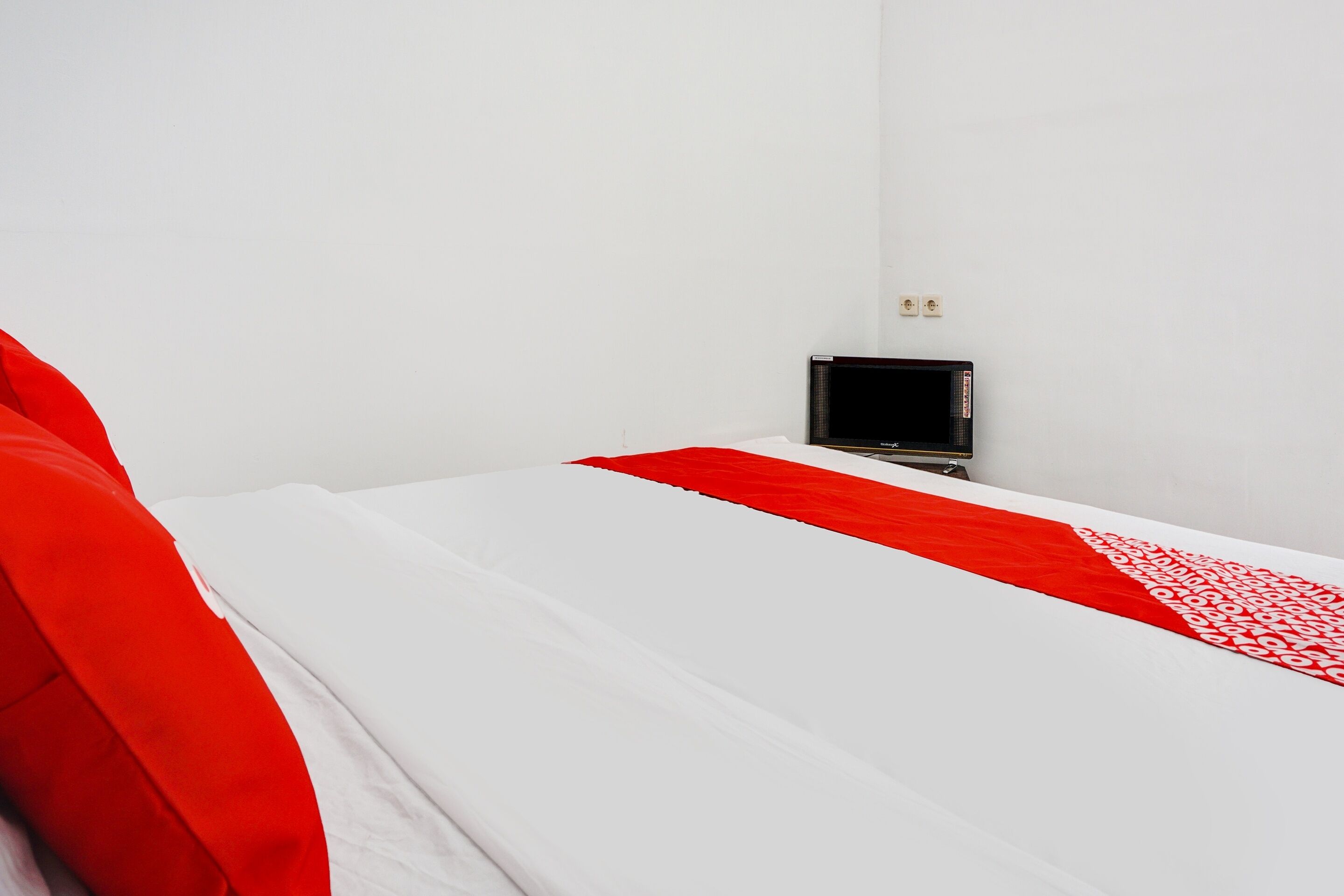 Una cama individual con una colcha blanca y roja, una mesita de noche con un objeto negro, y una pared blanca con dos enchufes.