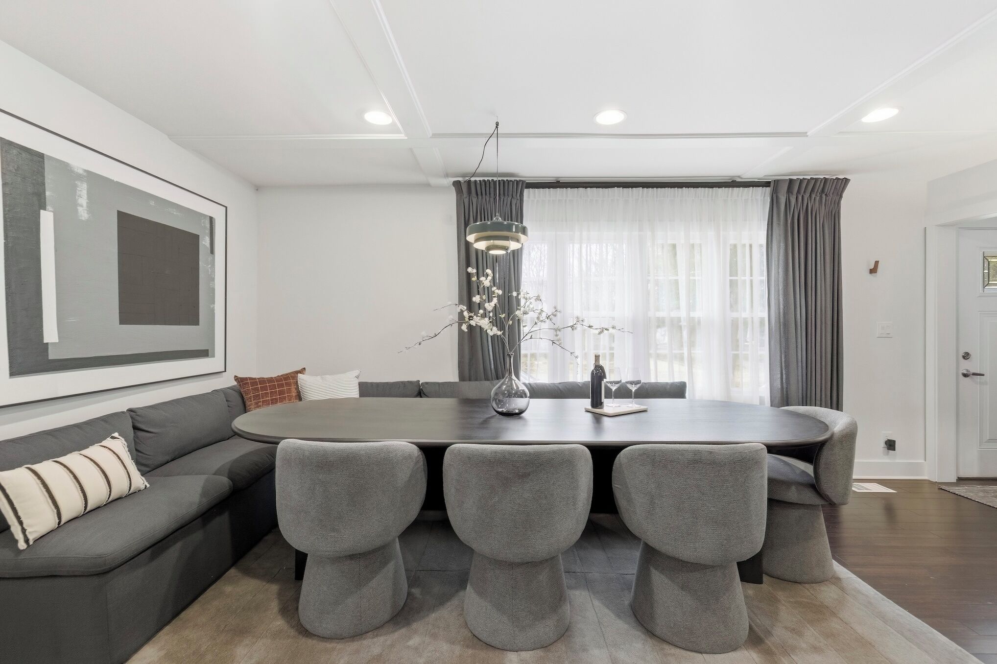 Un espace de salle à manger moderne avec une table ronde, des chaises recouvertes d’un tissu gris, un vase orné de branches et un luminaire suspendu.