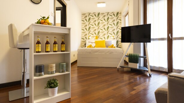 Un salon moderne avec un plancher en bois, un canapé blanc agrémenté de coussins jaunes, une étagère blanche où sont disposées des bouteilles, et un téléviseur à écran plat posé sur un meuble.