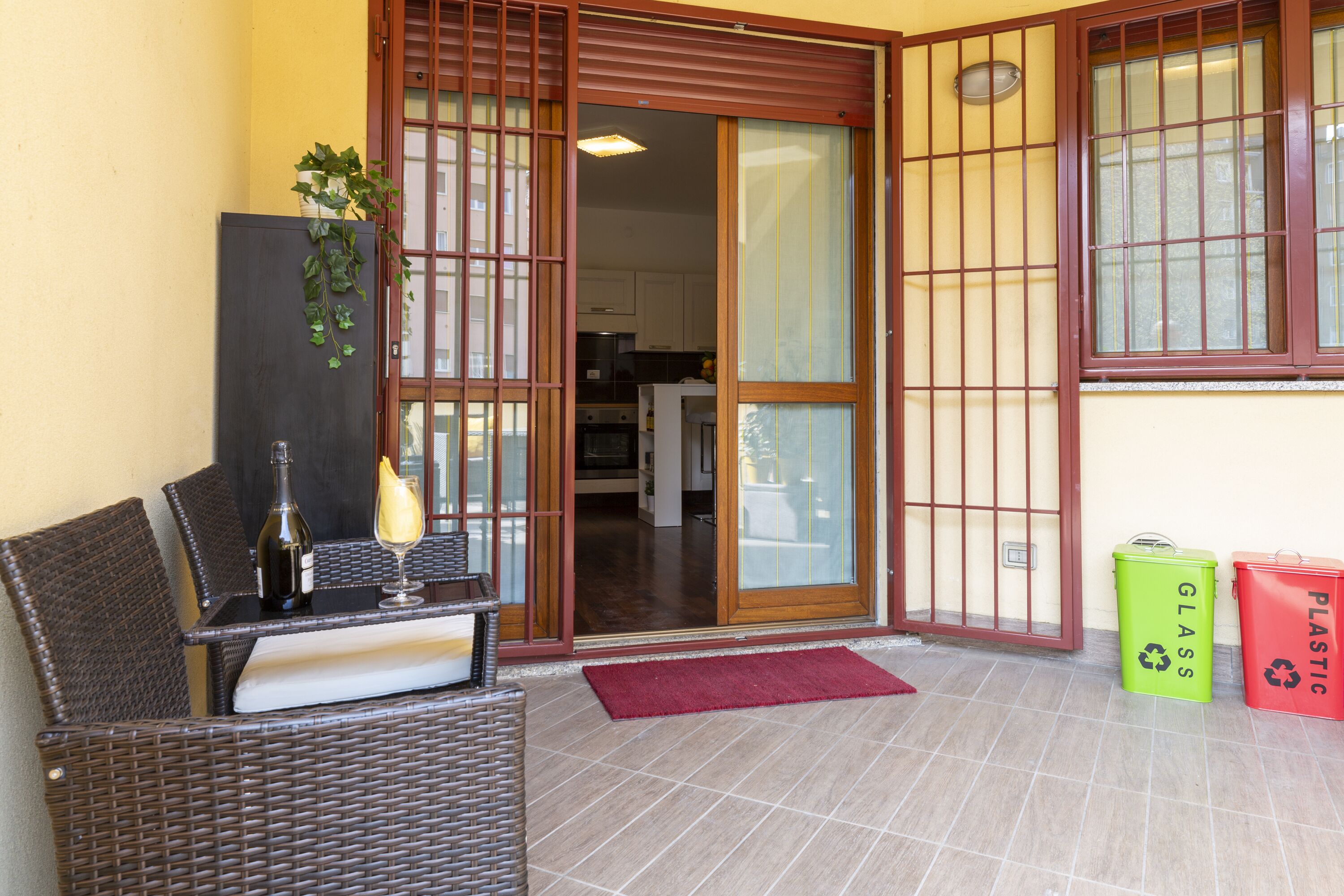 A room with a wicker chair, a bottle, and a glass on a table, a sliding door, and two trash bins labeled "glass" and "plastic".