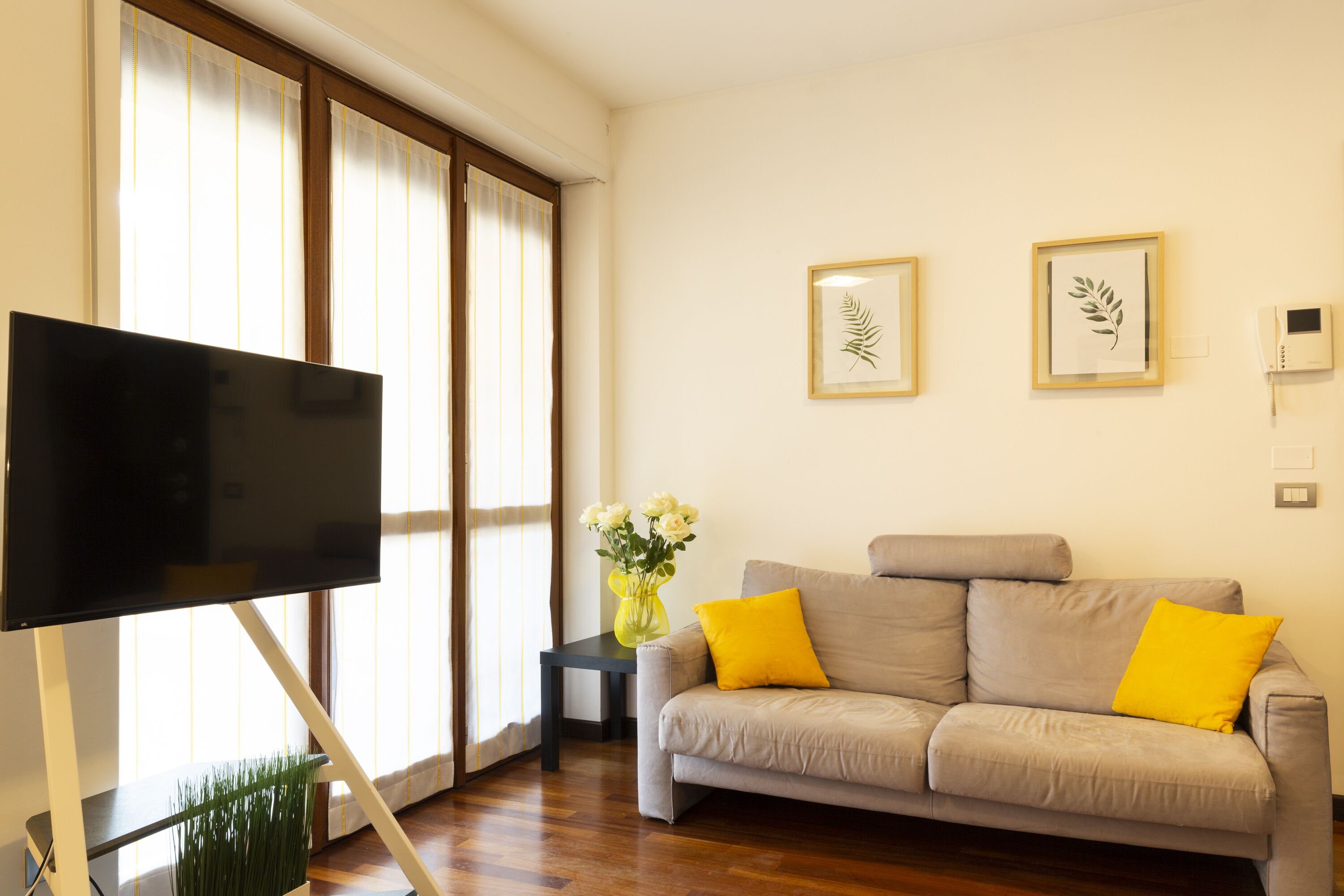 Una sala de estar con un sofá gris, cojines amarillos, un televisor de pantalla plana, una mesita con un jarrón de flores y arte con hojas enmarcadas en la pared.