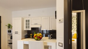 Une cuisine moderne avec des meubles blancs, un îlot central agrémenté d’un panier de fruits, et une vue sur une chambre à coucher avec un lit et des coussins jaunes.