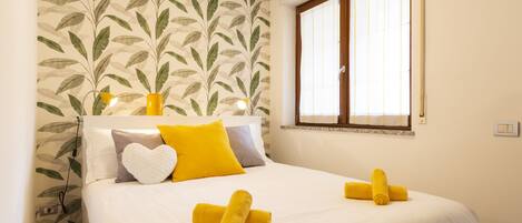 A neatly made bed with white bedding, a heart-shaped pillow, and two rolled towels.