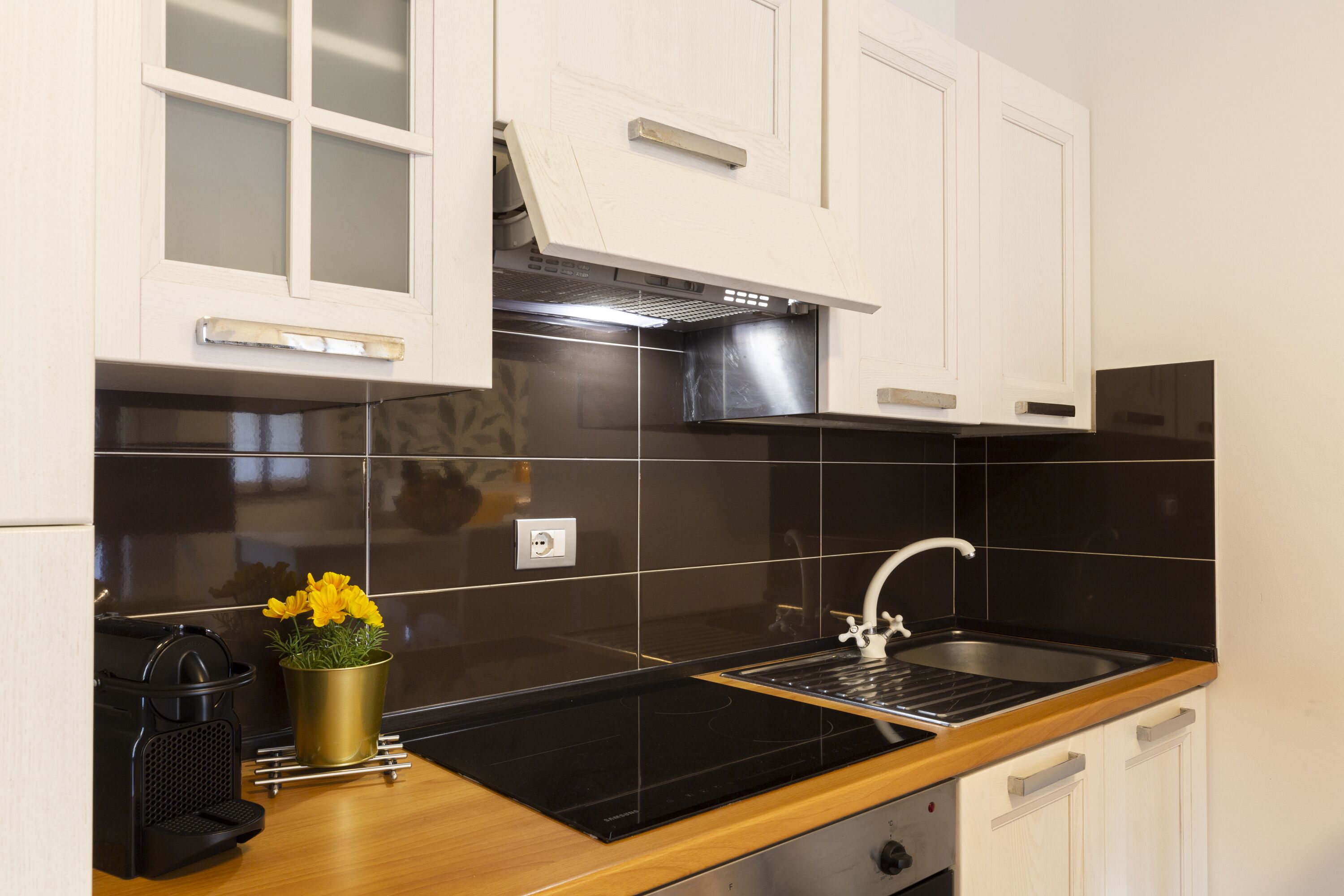 A modern kitchen with white cabinets, a black backsplash, a stainless steel range hood, a black induction cooktop, a wooden countertop, and a sink with a faucet.