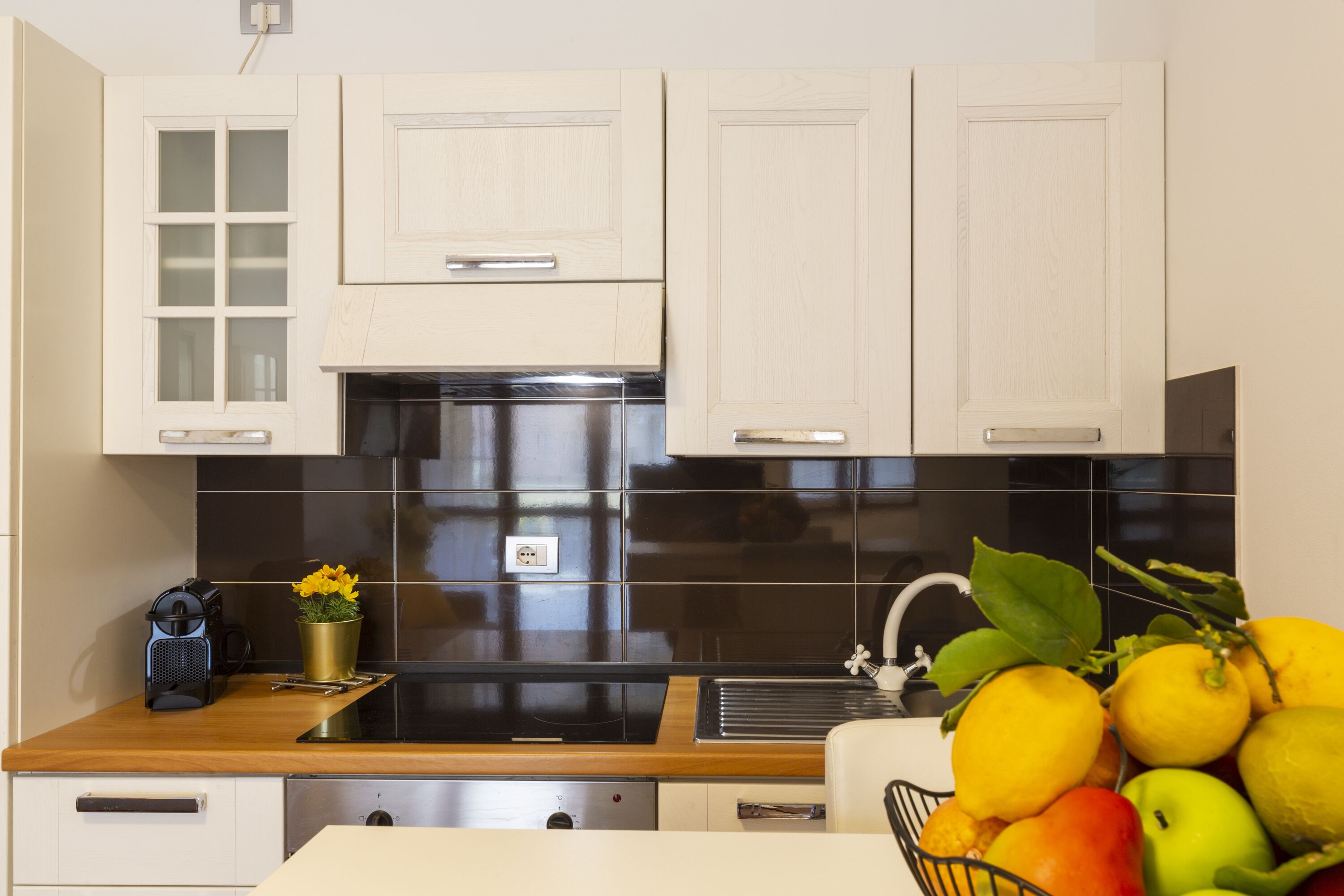 Una cocina con armarios blancos, salpicadero de azulejos negros, electrodomésticos de acero inoxidable y un frutero sobre la encimera.